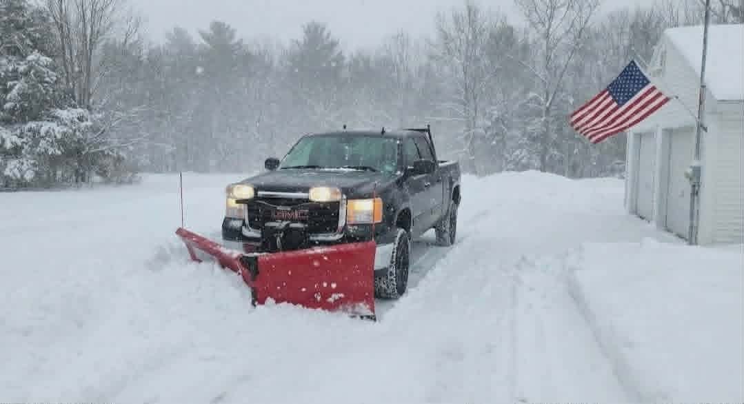 Black pickup truck with red snow plow clearing snowy road, American flag visible in background